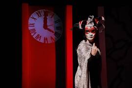 A photo from a scene of Masque of the Red Death. The character stands menacingly with a blood red jester mask and a grey cloak, ready to snap as the white clock on the blood red background chimes